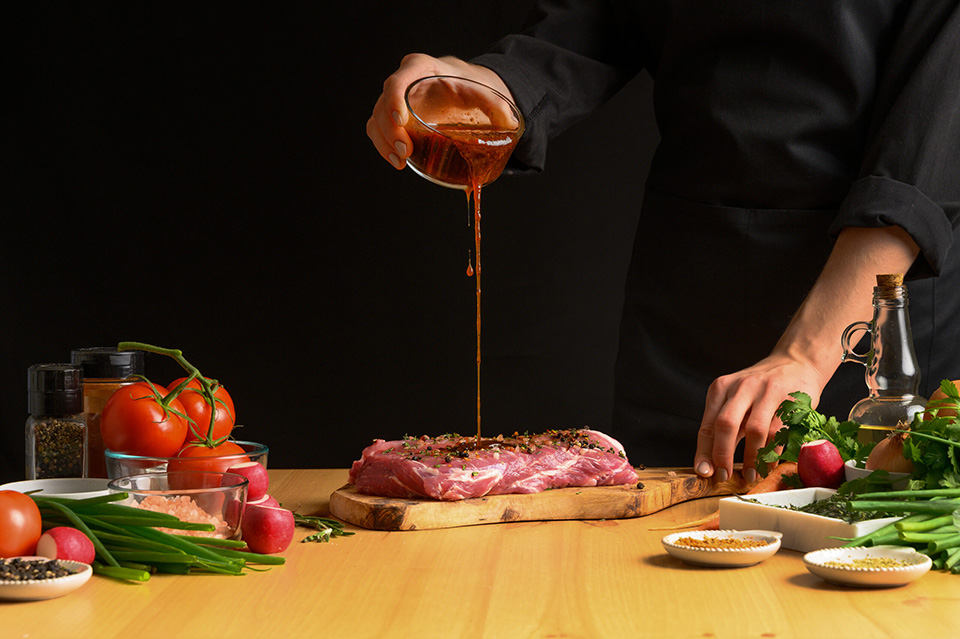 Chef cooks pork steak. pouring sauce, marinade on a background with vegetables. Recipe book, cooking, restaurant business