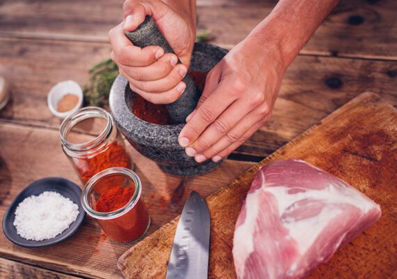 High angle shot of a herbs and spices being ground up in a stone mortar and pestle to make a dry rub seasoning for a piece of qulity raw pork lying alongside