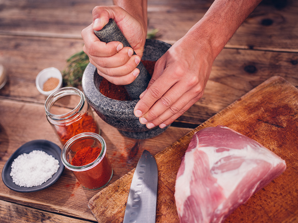 High angle shot of a herbs and spices being ground up in a stone mortar and pestle to make a dry rub seasoning for a piece of qulity raw pork lying alongside